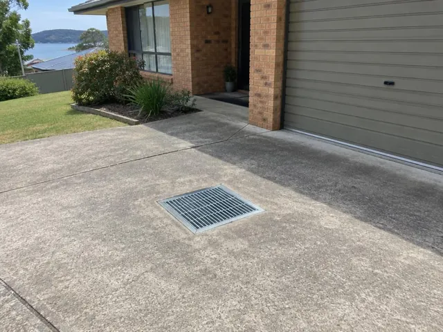Concrete driveway with a square outdoor drain grate beside a brick house in Buttaba